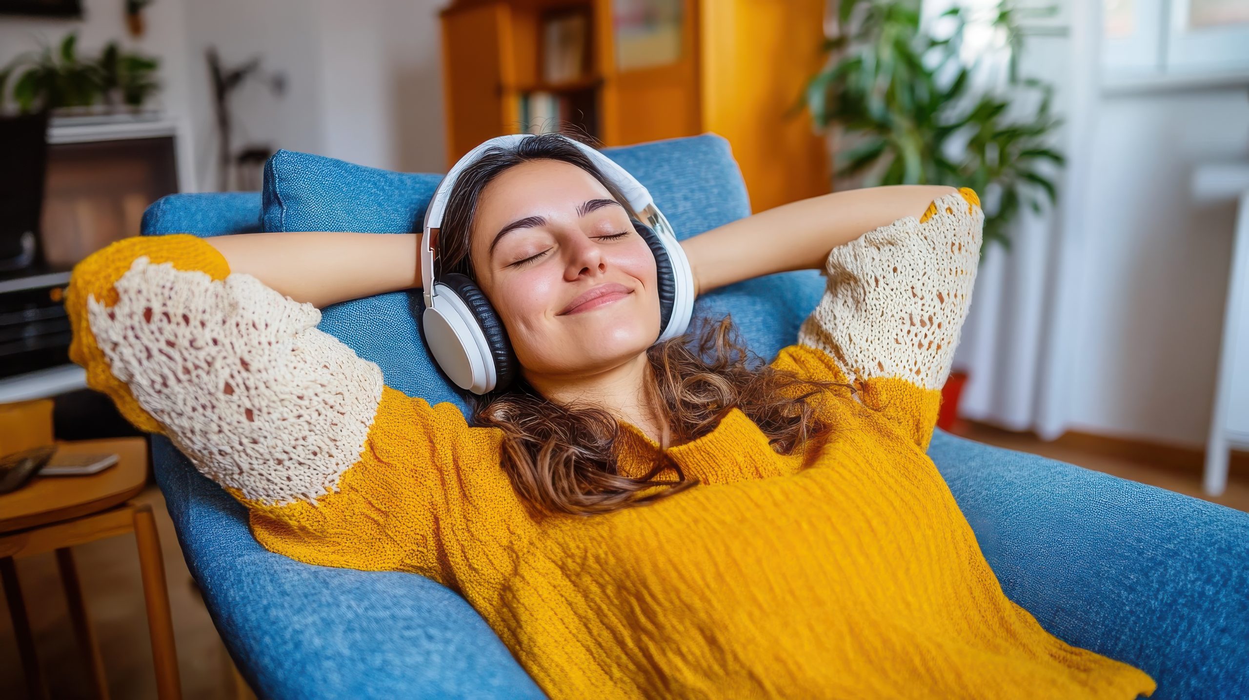 A woman in a crochet-detailed yellow sweater is stretched out on a blue chair, listening blissfully to music in a sunlit and stylish living room setting.